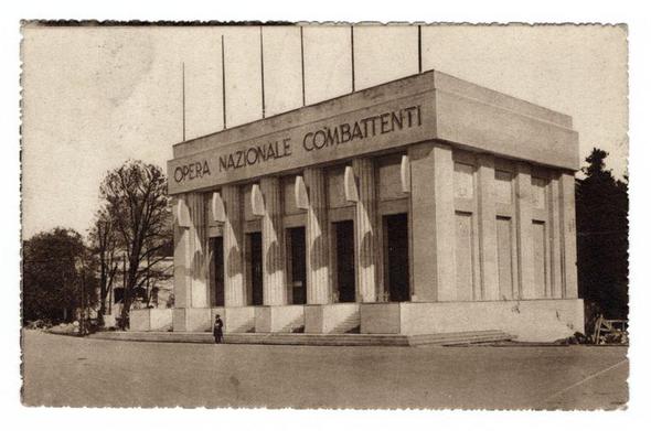 The image depicts a historical photograph of the Opera Nazionale Combattenti building, which is labeled as such in large letters on the facade. The structure has a strong neoclassical architectural influence with its use of columns and symmetrical design elements. It appears to be an older sepia-toned photo, suggesting it may have been taken around 1928, aligning with the date mentioned in the image source link provided (the Operazione Nazionale Combattenti exposition was held in Turin that year). The building is situated on a street lined with trees and bare-branched winter foliage. A solitary figure stands at some distance from the entrance of the opera house, adding to the historical ambiance of the scene. There are no visible modern elements or people dressed in contemporary clothing, emphasizing its vintage context.