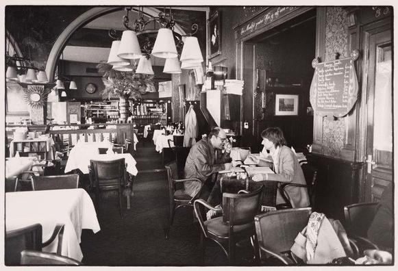 The image is a black and white photograph depicting an interior scene of what appears to be a restaurant. In the foreground, we see a couple seated at one of several tables draped with tablecloths, engaged in conversation over drinks or meals that are not fully visible on their plates. The man wears glasses and both individuals seem absorbed in discussion.

The setting is vintage, suggested by the design elements such as chandeliers, wall-mounted lamps, framed artwork, and a large mirror reflecting part of the dining area. There's an old-fashioned clock on one side and what looks like a menu board with handwritten text visible near the entrance or doorway to another section of the establishment.

The composition captures the ambiance of a bygone era restaurant, complete with patrons enjoying their time in this nostalgic environment. The photo is well-composed with depth, inviting viewers into both foreground activity and glimpses of other diners in the background.