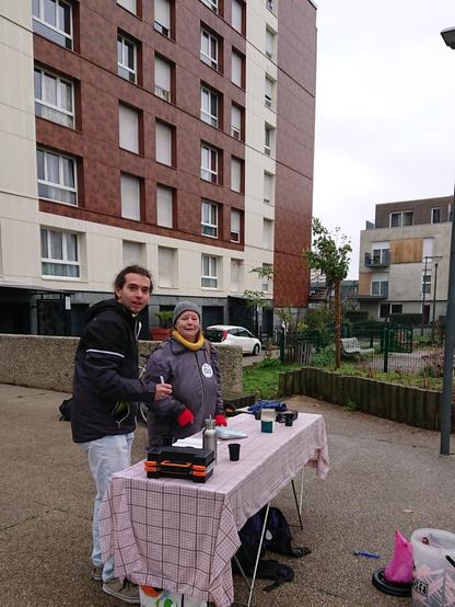 Jeune homme et femme bien couverts surpris par l'objectif en train de discuter à une table avec du café, des mallettes et une lampe portable, sur une place piétonne entre jardin et immeuble rénové.