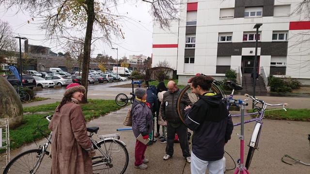 2 femmes, une fillette, 2 hommes et un garçon réparent ou discutent, leurs vélos à la main ou sur pieds d'atelier, sur une place piétonne non loin d'un grand œuf en béton et un grand bâtiment en bois.