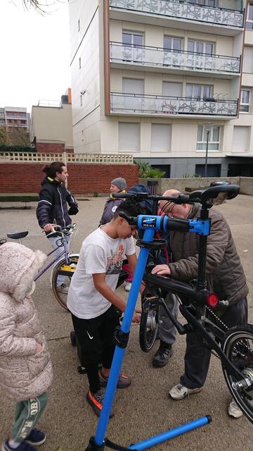 Un garçon pas frileux et un homme réparent une crevaison sur un VAE pliant. Une fillette les observe, tandis qu'un jeune homme tenant son vélo d'une main discute avec une bénévole de l'association.