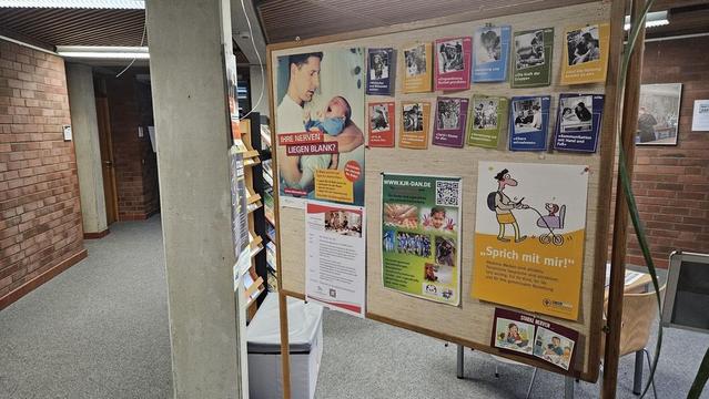 Plakatwand mit farbigen Infografiken und Texten, u.a. "Ihre Nerven" und "Sprich mit mir". Im Hintergrund ein heller Raum mit Stühlen.