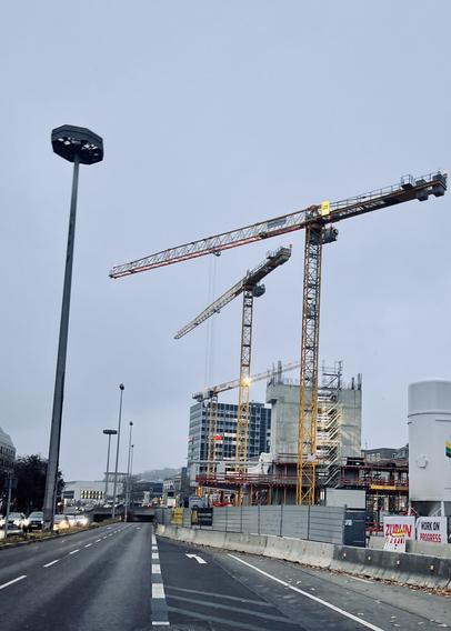 Große Baustelle neben einer mehrspurigen Straße. Gelbe Kräne ragen in den grauen Himmel, am Rand stehen helle Baucontainer und im Hintergrund sind die Rohbauten zu sehen.