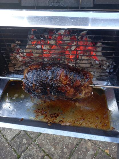 Ein gut gefärbter Spießbraten auf einem gußeisernen Holzkohlegrill, bei dem man hinter dem Braten die glühenden Kohlen sieht.