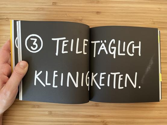 Innenansicht des Buches. Eine Doppelseite mit schwarzem Untergrund und weißer Schrift. Dort steht in Großbuchstaben: Teile täglich Kleinigkeiten.