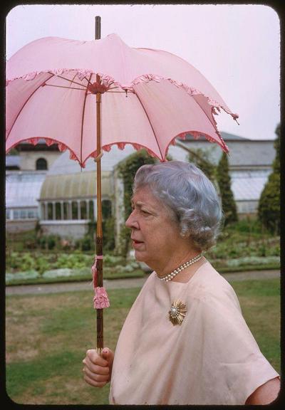 A woman is standing in the garden holding up an umbrella above her head and looking away from it, with a greenhouse visible in the background. She has silver hair tied back and wears pearl jewelry including a necklace around her neck and pin on her chest as well as gold tone flower brooches. The pinkish-pink umbrella she holds is frilly at edges and looks ornate.