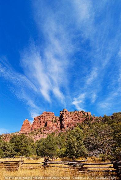 A layered red rock hill rises from a piñon forest. In the foreground a weathered wood rail fence, an old corral, stands in long sere grass. Above is an expanse of blue sky with thin white clouds seeming to radiate from the hilltop.
©BosqueBill.com