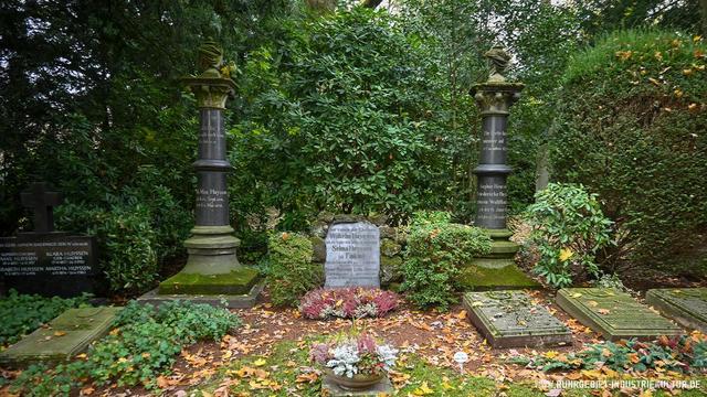 Historische Familiengrabstätte auf einem bewaldeten Friedhof. In der Mitte ein weißer Grabstein, flankiert von zwei dunkelgrauen Säulen mit Urnen. Im Vordergrund Herbstlaub und Bepflanzung mit Heidekraut. Dichtes Grün im Hintergrund.