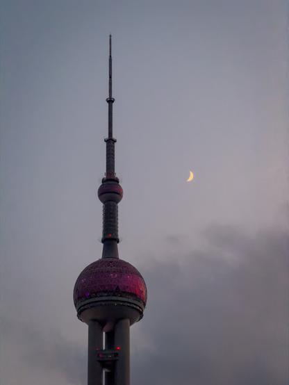 A twilight view of the Oriental Pearl Tower in Shanghai, with its illuminated magenta sphere standing out against a soft grey-blue sky. A thin crescent moon glows to the right of the tower, adding a delicate focal point above the evening clouds.