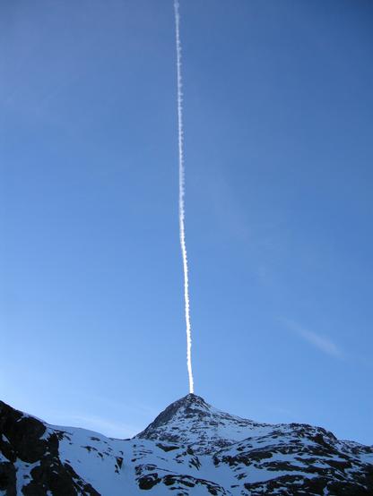 Ein Berg im Winter, blauer Himmel, ein Ein Kondensstreifen, der genau auf dem Berggipfel ankommt bzw. von ihm auszugehen scheint und das Bild senkrecht durchschneidet.