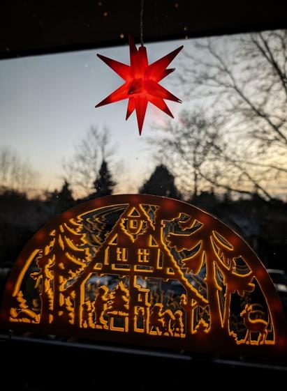 Ein orangefarbenes, ausgeschnittenes Fensterbild hängt vor einem Hintergrund aus Bäumen, der in die Dämmerung übergeht. Das Bild ist halbkreisförmig und zeigt eine Szene mit Häusern, Bäumen und Figuren. Darüber befindet sich ein Stern mit mehreren Spitzen, der durchscheinend ist und durch das Licht beleuchtet wird. Der Schwibbogen ist ausgeschnitten, sodass der Hintergrund durch die Szene hindurchscheint.