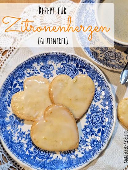 Holztisch, darauf ein rundes, weißes Deckchen mit gehäkeltem Spitzenrand. Auf dem Deckchen ein weißer Porzellanteller mit blauem, floralem Muster (Serie Burgenland), daneben eine passendes Kaffeetasse samt Untertasse und Löffelchen. In der Tasse Kaffee mit heller Crema. Auf dem Teller drei hellgelbgoldene, mit hellem Zuckerguss glasierte Zitronenkekse. Teils sind noch Stücke von Zitronenschale in der Glasur sichtbar. Neben dem Teller liegen weitere Kekse auf dem Deckchen. 
Oben am Bildrand ist ein halbtransparentes, weißes Feld. Darin steht "Rezept für Zitronenherzen [glutenfrei]".