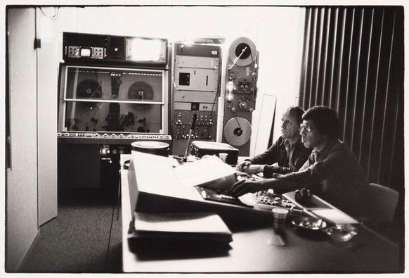 The image is a black and white photograph depicting two individuals in what appears to be an audio recording studio. The person on the left, presumed male due to hairstyle and attire, sits with his legs folded underneath him and hands resting on papers placed atop a large piece of paper or document lying across a table. His attention seems focused towards something off-camera.

The individual on the right is engaged in operating audio equipment situated behind them; this includes various pieces such as tape recorders, consoles, dials, switches, and other control panels indicative of professional recording gear from an earlier era given their design. The operator's hands are actively manipulating knobs or levers on one of these devices.

Behind both individuals stands a large soundproofed wall typical in audio studios to minimize echo and noise interference during recordings. In front of them is a window, which reveals daylight illuminating the interior space they occupy. A reflection can be seen on an adjacent glass surface indicating overhead lighting within the room.

Several items are scattered across what appears to be a worktable: papers, glasses with beverages or possibly recording equipment accessories like cables and perhaps small boxes for audio components. The overall setting suggests collaboration in producing sound recordings, potentially related to radio broadcasting given the context provided by the caption [...]