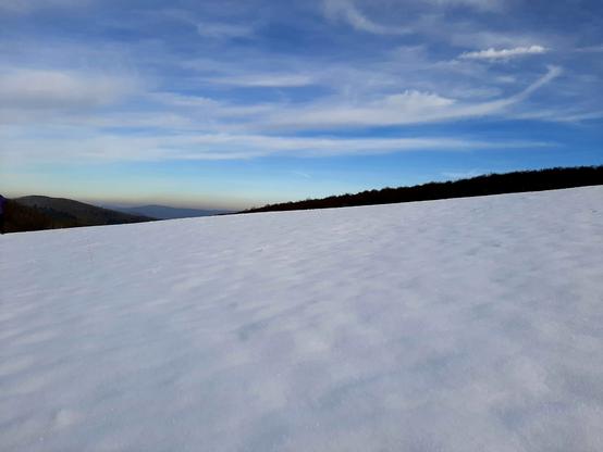 Eine weite leicht abfalkende Schneefläche, im Hintergrund schwarze Silhouetten von Wäldern, der Himmel ist blau mit langgezogenen und teilweise leicht gebogenen Wolken. Am Horizont schimmert ein Streifen leicht orangefarben.