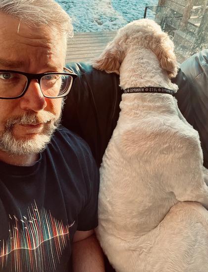 A person with glasses and a beard is sitting on a couch next to a dog looking out the window.