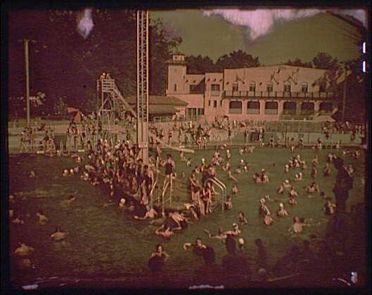 The image depicts a crowded outdoor swimming pool during the summer months, with numerous people enjoying various activities in and around the water. The scene shows individuals engaged in play on swings above the pool, while others swim or relax in the water below.

In addition to swimmers, there are groups of children playing games like duck-duck-goose near the edges of the pool, creating a lively atmosphere. Several people can be seen sitting along the grassy areas surrounding the swimming area, either supervising their children or simply watching the activities unfold.

A large building with multiple levels and balconies is visible in the background behind the trees, likely serving as changing facilities or part of the park's amenities for visitors. The overall setting suggests a popular recreational spot during this era, potentially from around 1920 to 1950 based on its visual characteristics.

The image captures the essence of outdoor leisure and community gatherings in parks like Glen Echo amusement park, reflecting how people interacted with public spaces for entertainment and socialization. The historical context hints at a time when such communal activities played an essential role in daily life, especially during warm weather months.