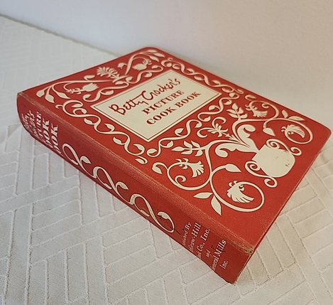 Color image of the 1950s-era Betty Crocker Picture Cookbook, at one time in virtually every American kitchen. Image shows a three-ring binder with a red and white cover in fanciful designs of white on red, with the book's title.