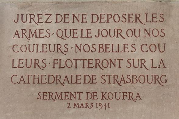 Gros plan sur une plaque de pierre claire, portant une inscription en capitales de couleur rouge-brun. Le texte est : "JUREZ DE NE DÉPOSER LES ARMES QUE LE JOUR OÙ NOS COULEURS NOS BELLES COULEURS FLOTTERONT SUR LA CATHÉDRALE DE STRASBOURG" suivi de "SERMENT DE KOUFRA" et "2 MARS 1941" en plus petit au bas.