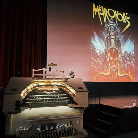 A Mighty Wurlitzer pipe organ sits off to the side of a grand movie theatre screen with the poster for 1927's Metropolis shining brightly upon it, towering above the theatre stage.