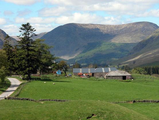 Glen Clova. The image shows the broad glen in the foreground, narrowing in the distance as it encounters mountains. Which occupy much of the upper half of the frame, leaving a gap for the glen in the upper left beyond trees. In the right middle of the frame is a group of farm buildings with grassy fields this side of them. The scene is in sunlight.