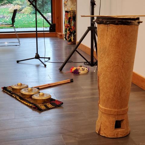 A tall, vertical, cylindrical wooden drum with a stretched animal skin top and a small square opening near the base, accompanied by three bronze gongs on a decorative cloth, two bamboo mallets, and a drumstick resting on top, set up for a performance. A tambourine is in the background, on the floor.