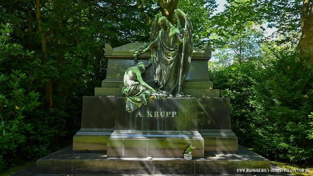 Imposantes Grabdenkmal mit bronzegrüner Engelskulptur, die eine trauernde Figur tröstet. Auf dem massiven Steinsockel die Aufschrift "A. KRUPP". Das Monument steht in üppigem Grün, umgeben von Rhododendren.