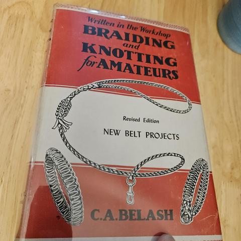 Written in the Workshop
Braiding amd Knotting for Ameteurs

Revised Edition
New Belt Projects
C.A. BELASH

A red and white book, Red band just under haflways down, then white, then a red bottom to abojt a quarter way up. 
There are 2 bracelets and a leash item drawn on it also, showcasing what may come in the material.