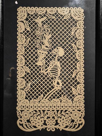 Intricate beige papel picado showing a kneeling skeleton offering a cup with flowers and birds against a lattice background.