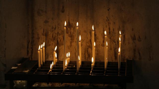 In einem dunklen Kirchenraum brennen einige schlanke weiße Kerzen auf einem schwarzen Metallgitter. Ein paar Kerzen sind schon fast heruntergebrannt, ein paar sind gerade erst angezündet worden.
//
In a dark church room, some slim white candles are burning on a black metal grate. Some candles have already almost burned down, some have just recently been lighted.
