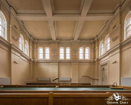 An abandoned courthouse, photographed by Obsidian Urbex. The room features arched windows, wooden railing, and faded beige paint.