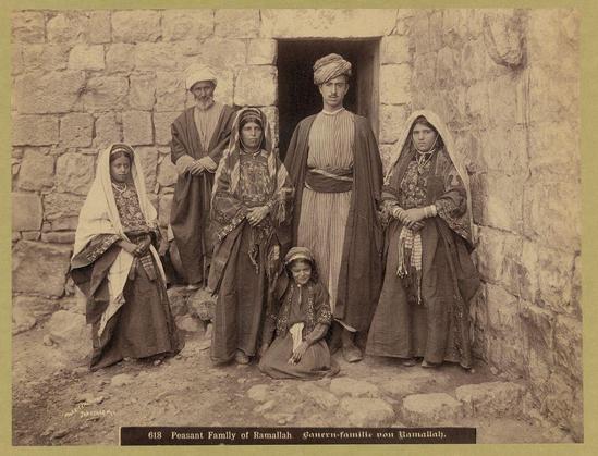 The image depicts a family portrait of six individuals, presumably from the Peasant Family of Ramallah. The setting appears to be an old stone building with a rustic ambiance. There are three men and two women standing behind them, along with one young girl sitting on her knees in front.

Starting from left to right: The first woman is dressed in traditional attire consisting of a long dress with intricate patterns, a head covering, and she carries something draped over her shoulder. Next to her stands an older man wearing a turban-style hat, a coat, and glasses, exuding an air of authority or respectability.

The third individual from the left appears to be younger than the others; his attire is more casual compared to those beside him - he wears a simple robe with stripes on it. Behind them all stands another woman adorned in heavy jewelry such as necklaces and bracelets, her expression somber but dignified.

To complete this portrait are two young girls: one sitting in front of the group appears quite small for their age; she's also wearing traditional clothing including a head covering that matches others' attire around here. The final girl is dressed similarly to those standing behind—her outfit decorated with vibrant colors and intricate embroidery suggestive of cultural significance.

The overall composition suggests this photograph was taken during an earlier period, possibly between 1900-1910 as indicated by the [...]