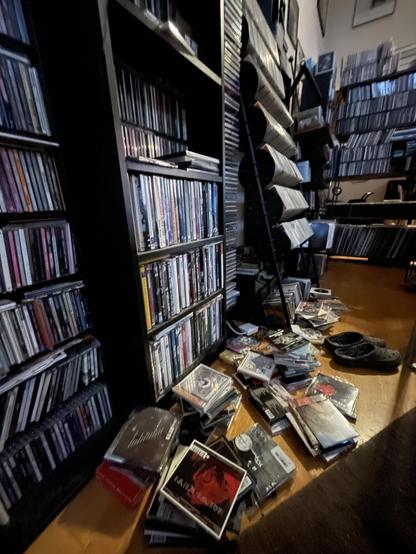 Short messy stacks of CDs are strewn about the floor in front of a wall lined with CD-packed shelves.