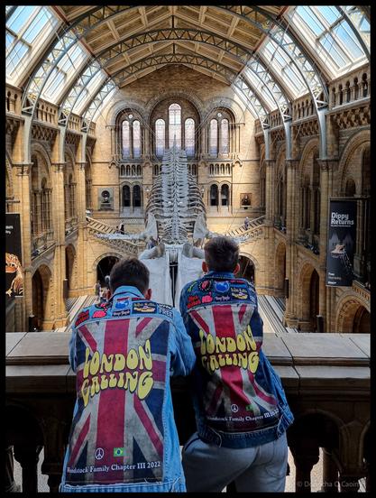 Photograph taken from behind two visitors wearing denim jackets printed with "LONDON CALLING" in yellow text over Union Jack designs and "Woodstock Family Chapter III 2022" patches, standing at a railing overlooking the main hall of the Natural History Museum London with a large skeleton mounted centrally beneath a soaring vaulted glass and timber roof with ornate stone columns and Gothic arches.