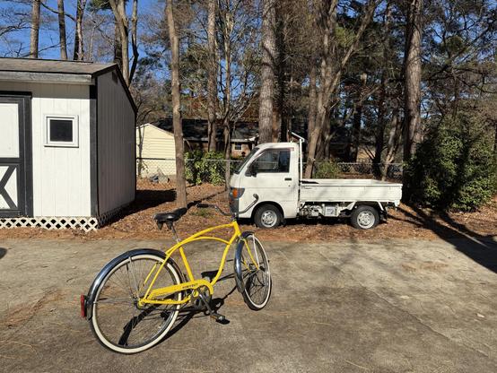 Yellow vintage bicycle, a Schwinn Heavy Duti with a curvy yellow frame and white wall tires, parked in front of a little white Japanese kei truck.