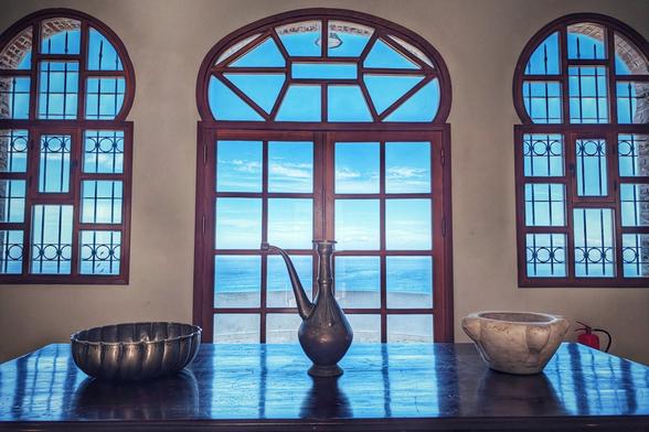A still life composition featuring a dark, highly polished wooden table set against a wall with three arched windows overlooking a bright blue sea. The central window is a large wooden-framed double door with a Moorish-style arch above. On the table, from left to right, are a hammered metal bowl, a slender antique metal jug (dallah or ewer), and a rough, light-coloured stone bowl. The light streams in, reflecting strongly on the table's surface, highlighting the deep blue of the sky and ocean outside.