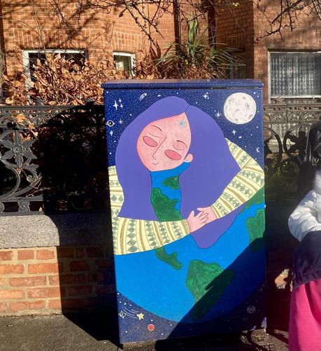 A lovely calming mural of a woman with her arms around the world on a traffic box in front of a terrace of red brick houses. There is a glimpse of B to the right, who is wearing her snow Queen coat and is covered with a pink GaryB blanket