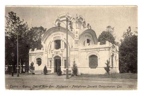 The image depicts a vintage black-and-white photograph of an ornate building known as the "Padiglione Società Consumatori Gas" from the 1902 International Exhibition held in Turin, Italy. This particular pavilion was designed by architect Raffaello Pelaia and is notable for its decorative elements inspired by baroque architecture. The structure features a central entrance with an archway supported by two columns topped with ornate capitals. Above this entry, there are multiple levels of decorative molding and sculptures that add to the building's grandeur.

The pavilion has three visible arched windows on the upper level above the main facade, adorned with intricate designs. On either side of these central features stand slender lamp posts or streetlights, adding symmetry to the composition. The photograph appears aged, as indicated by its monochromatic color scheme and slight discoloration around edges.

Trees line up along a path in front of the building, suggesting it was located within an urban park setting during that era. A caption at the bottom left corner reads "Torino - Espos. Int. d'Arte Dec. Moderna - Padiglione Società Consumatori Gas (12)," which translates to "Turin - International Art Exhibition of Modern Decorative Industry - Pavilion Consumers Gas Society." The overall impression is that of a historical building, preserved as an example of early 20th-century industrial architecture with artistic flair.