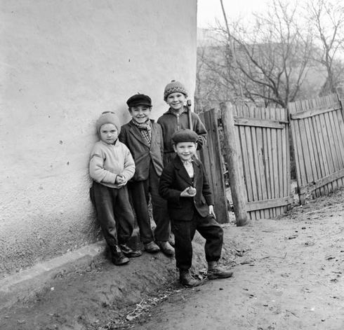 A black and white photo of  four children smiling while looking at the camera while standing beside a wall in a rural setting. They are dressed in hats and jackets suggesting cooler weather.  One boy, who looks like the oldest of the group, is carrying a small axe over his shoulder.