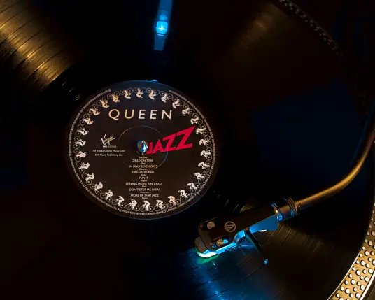 Die LP "Queen - Jazz" liegt auf dem Plattenteller, der Tonarm befindet sich im letzten Track des Albums. Der Tonarm wird von einem kleinen, bläulichen Licht beleuchtet, das sich in der schwarzen Vinyl spiegelt.