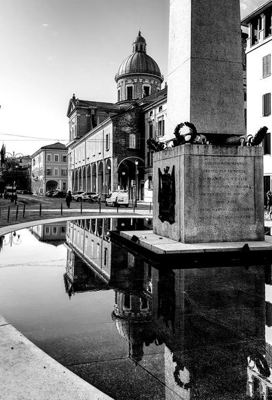 Foto verticale in bianco e nero di Piazza Gioberti a Reggio Emilia. In primo piano câĂš la base di un monumento in pietra, con iscrizioni e corone, riflessa nella vasca dâacqua davanti. Nellâacqua si specchiano anche la cupola e la facciata della chiesa e degli edifici porticati sullo sfondo. Si vedono alcune auto parcheggiate e una persona che cammina nella piazza, sotto un cielo chiaro.
