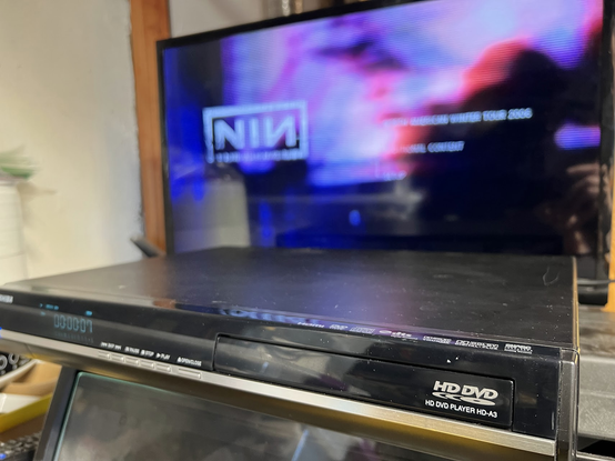 The top-level menu for Nine Inch Nails Beside You In Time with the NIN logo in white on heavily blurred concert imagery animated in the background.