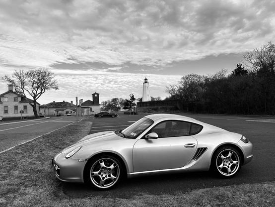 A silvery Porsche Cayman is shown in the foreground in front of a lighthouse in the distance and slightly stormy looking clouds.
The image is in B&W which is not very different from the in real life look at the time