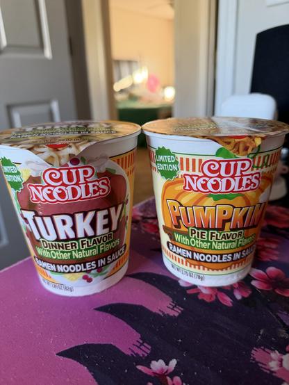 Two cylindrical containers of instant noodles are sitting side-by-side on a patterned surface. Both containers are orange and gold and have a "Cup Noodles" logo. The container on the left features an image of turkey and reads, "LIMITED EDITION" at the top and "TURKEY DINNER FLAVOR" in large letters, followed by "With Other Natural Flavors" and "RAMEN NOODLES IN SAUCE" underneath. The container on the right has an image of pumpkin pie and reads, "LIMITED EDITION" at the top and "PUMPKIN PIE FLAVOR" in large letters, followed by "With Other Natural Flavors" and "RAMEN NOODLES IN SAUCE" underneath. Both containers are sealed with a peel-back lid, and have a net weight of 2.05 oz (58g) printed on the bottom. The patterned surface appears to be a tablecloth with pink and yellow flowers.
