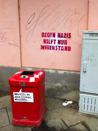 Ein roter Mülleimer steht auf einem Gehweg vor einer rosafarbenen Wand. Auf dem Mülleimer klebt ein Aufkleber mit der Aufschrift „Wirf es in den Eimer, Kleines.“ Die Wand ist mit Graffiti und einem markanten, roten Schriftzug besprüht, der „Gegen Nazis hilft nur Widerstand“ lautet. Daneben sind Kritzeleien und ein Davidstern zu erkennen. Der Boden ist uneben und zeigt Spuren von Schmutz und Laub. Rechts neben dem Mülleimer steht ein grauer Verteilerkasten. Die Farben sind kontrastreich, das Rot des Mülleimers sticht hervor und zieht den Blick auf sich. Die Stimmung wirkt urban und rebellisch, vermittelt durch die politische Botschaft und die unordentliche Umgebung. [KI-generiert]