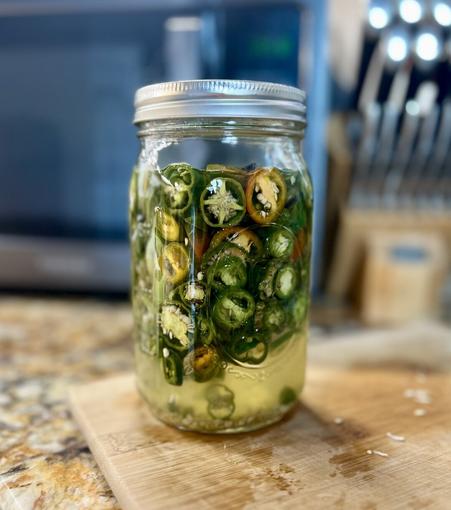 A glass mason jar filled with freshly pickled jalapeño, poblano, &amp; bell pepper slices, sitting on a wooden cutting board, with a blurred kitchen background.