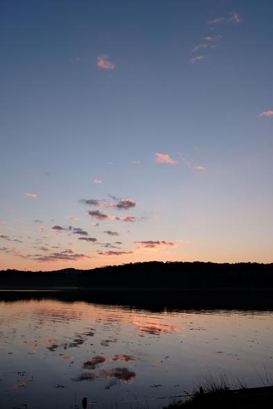 A lakeview at dawn as the sun paints the sky and clouds in orange over woods. The water reflects the clouds and orange color.