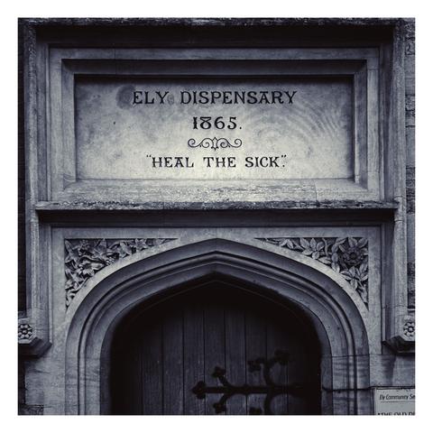 Square black and white photo of a stone doorway with a an inscription in the headstone saying ‘Ely Dispensary, 1865, “Heal the Sick”’ and a wooden door below