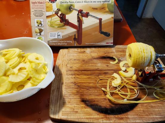A yellow, peeled apple attached to a metal arm is over a wooden board. The apple is still attached but is in slices and the peel lies on the board beneath. Behind is the box for the tool, called an Apple Mate 2, with a picture of the red and silver metal tool in use anf a claim that it "Quickly Pares, Cores & Slices in on easy motion!". On the left is a white bowl filled with sliced, peeled apples