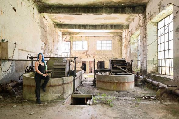 A woman with blue hair poses inside an abandoned industrial ruin with decaying equipment and peeling walls. Two large water wheels sit inside concrete tanks.