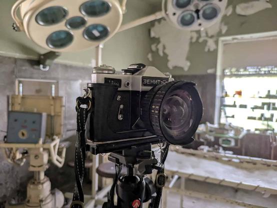 A vintage Zenit camera on a tripod sits inside an abandoned hospital room. Surgical lights and decaying walls indicate an urban exploration setting.